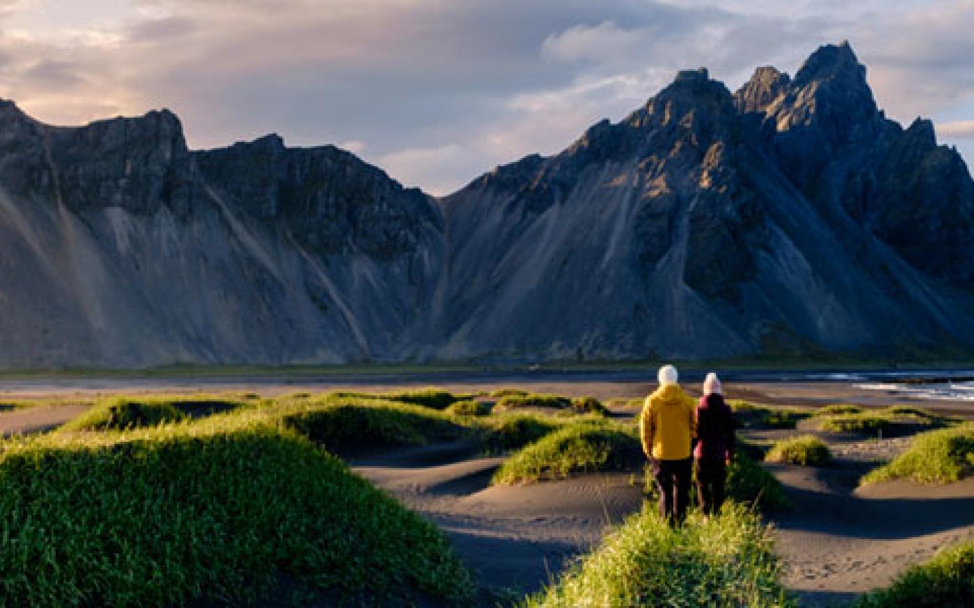 Two people walking through a grassy coastal landscape toward dramatic rocky mountains.