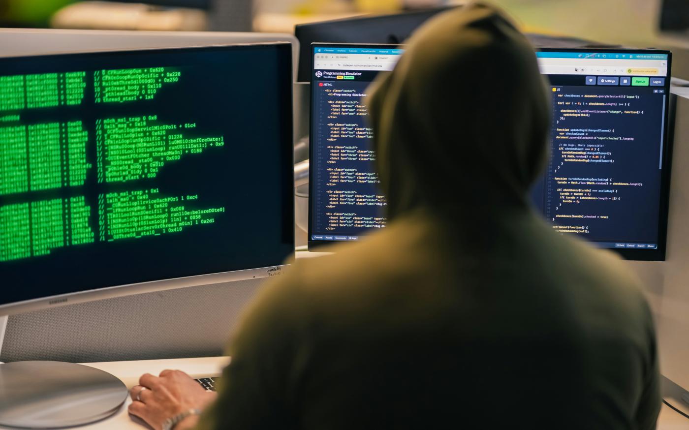 Hacker in hoodie working on multiple computer screens by Julio Lopez courtesy of Unsplash.