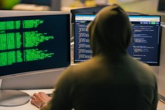 Hacker in hoodie working on multiple computer screens by Julio Lopez courtesy of Unsplash.