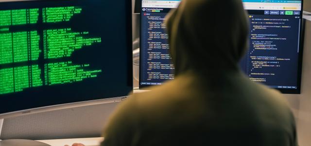 Hacker in hoodie working on multiple computer screens by Julio Lopez courtesy of Unsplash.